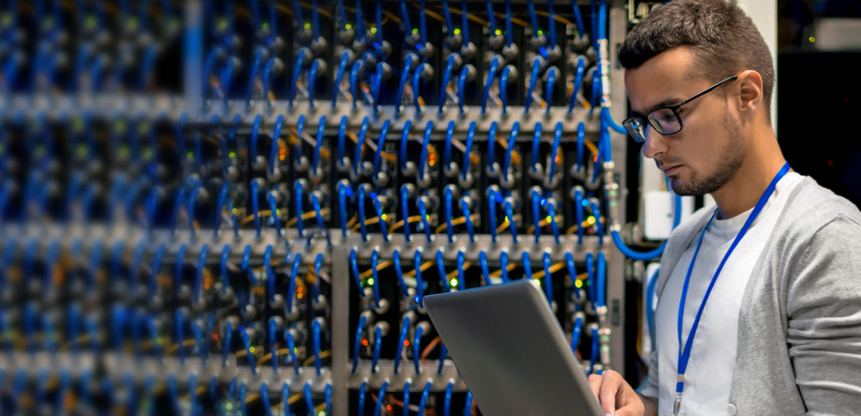 A man looking at a laptop in front of a wall of servers.