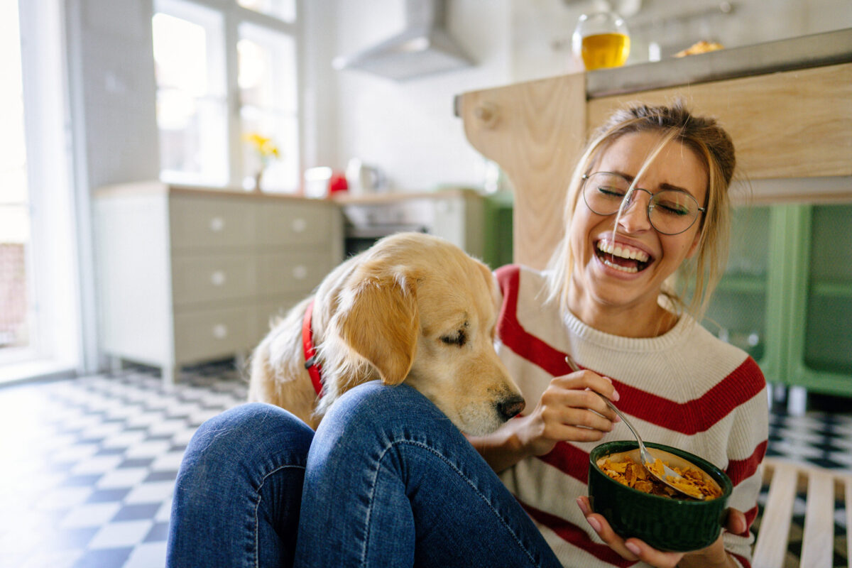 A woman laughing as her dog looks at food in her bowl.