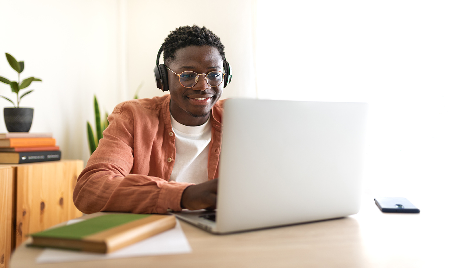 A smiling young man wearing glasses and headphones as he looks at a laptop.