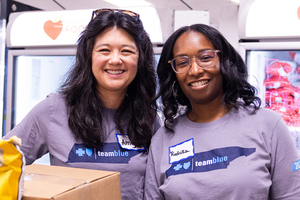 Two women in t-shirts standing next to boxes, engaged in a volunteer activity.